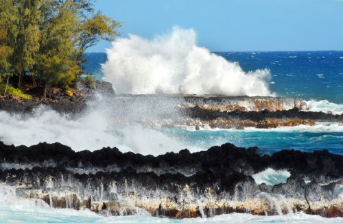 Waves crash against rocky shorelines, with lush trees in the background and a bright blue sky above.
