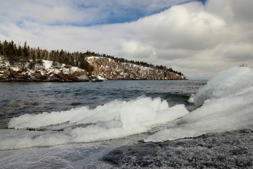 A winter landscape featuring icy rocks by a lake, with snow-covered cliffs and cloudy skies in the background.
