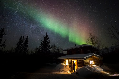 A person stands at the door of a cabin under a starry sky illuminated by the northern lights. Snow covers the ground.