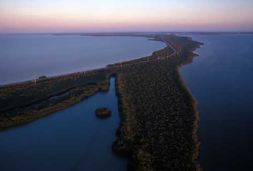 Aerial view of a coastal landscape with water, lush greenery, and a road lined with power poles at sunset.