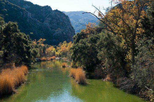 A serene river flows through a lush landscape with autumn foliage and rocky hills in the background.