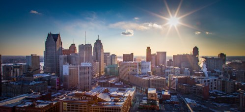 A panoramic view of a city skyline at sunset, with tall buildings and a bright sun shining in the background.