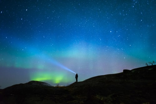 A silhouette of a person with a flashlight stands on a rocky landscape under a starry sky and colorful auroras.