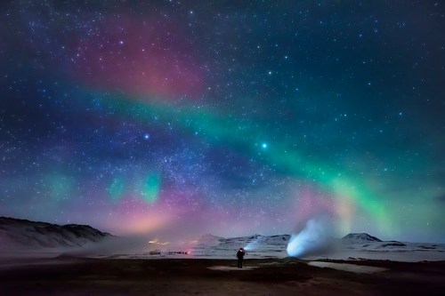 A person stands under a vibrant night sky filled with colorful auroras and stars, with mountains in the background.