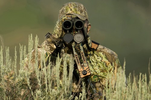 A camouflaged hunter using binoculars, surrounded by tall green vegetation.