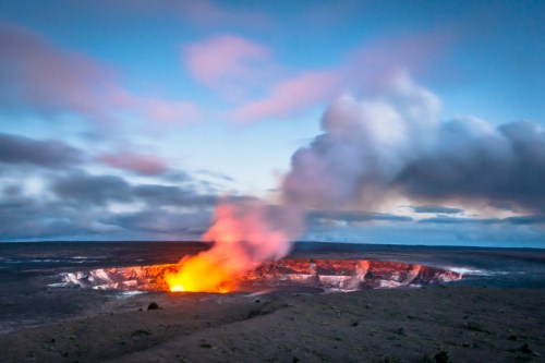 A volcanic crater emits bright orange lava and smoke against a twilight sky.