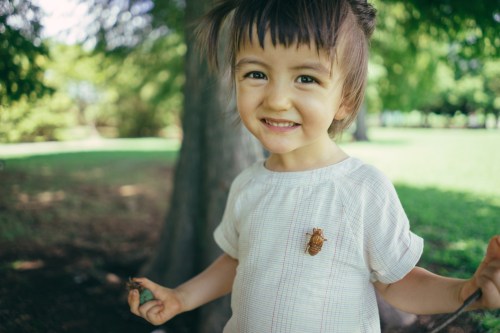 A smiling child in a light shirt stands outdoors, holding a twig, with a tree and green grass in the background.