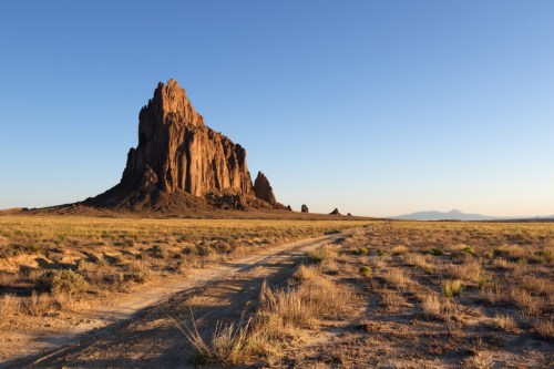 A towering rock formation rises above a desert landscape under a clear blue sky, with a dirt path leading towards it.