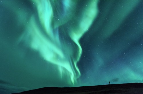 A person stands on a hill under a vibrant green aurora borealis lighting up the night sky filled with stars.