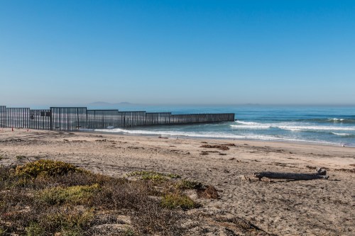 A coastal view of a border fence along the beach, with waves and clear blue skies in the background.