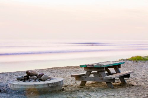 A picnic table beside a fire pit on a sandy beach, with calm waves and a pastel sky in the background.