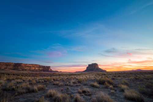 A vast desert landscape at sunset, featuring distant mesas and a colorful sky with soft clouds.