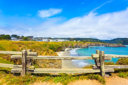 Scenic coastal view with a wooden fence, sandy beach, and vibrant blue water under a partly cloudy sky.