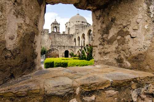 View through a stone archway showcasing a historic building with domes and lush greenery in the foreground.