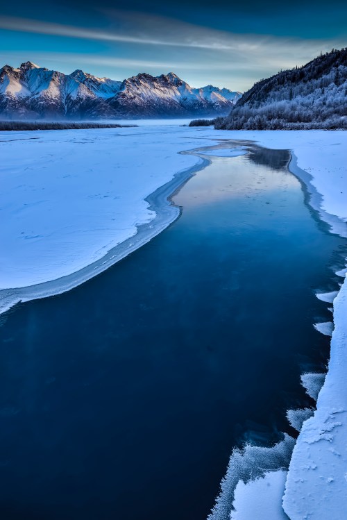A serene winter landscape featuring a frozen lake, snow-covered mountains, and a winding river reflecting the sky.