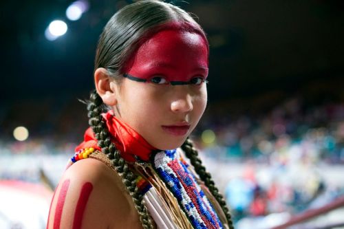 A young girl with traditional face paint and braided hair, wearing colorful beads, gazes confidently at the camera.