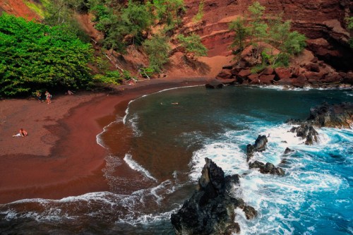 A scenic view of a red sand beach with rocky shores and turquoise waves, surrounded by lush greenery.