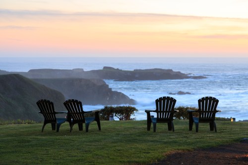 Four black Adirondack chairs facing a serene ocean view at sunset, with gentle waves and rocky coastline in the background.