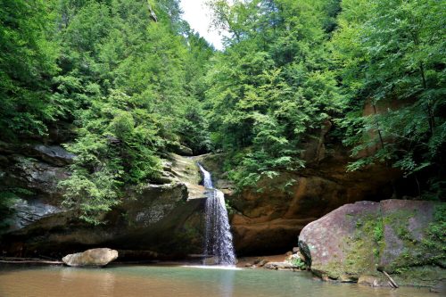 A serene waterfall cascades down rocky cliffs, surrounded by lush green trees and a calm, reflective pool below.