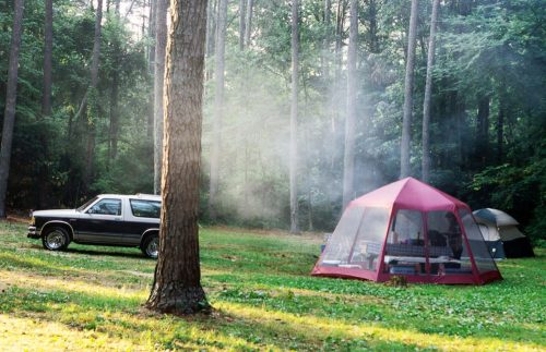 A campsite in a forest with a black SUV, a red tent, and a gray tent, surrounded by trees and light smoke.