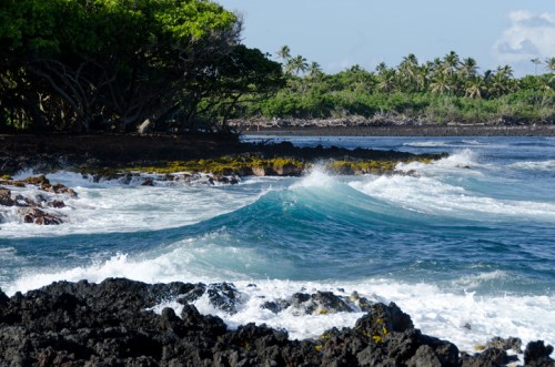 Waves crash against rocky shorelines, surrounded by lush greenery and palm trees under a clear blue sky.
