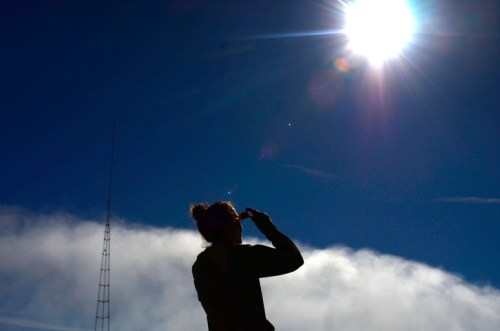 A silhouette of a person looking up at the bright sun against a clear blue sky with clouds below.