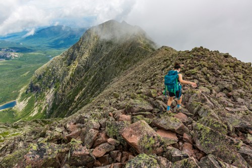 A hiker navigates rocky terrain on a mountain ridge, surrounded by clouds and lush green valleys below.