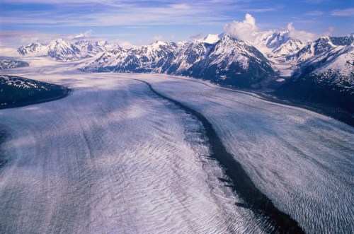 Aerial view of a vast glacier flowing through a mountainous landscape, surrounded by snow-capped peaks and blue skies.