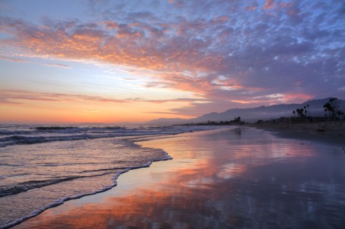 A serene beach at sunset, with colorful clouds reflecting on the wet sand and gentle waves lapping at the shore.