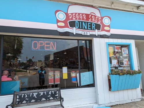 A colorful diner with a retro car sign, featuring an "OPEN" neon sign and a flower box under the window.