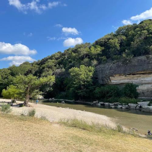A serene riverbank scene with lush greenery, rocky cliffs, and a clear blue sky dotted with clouds.