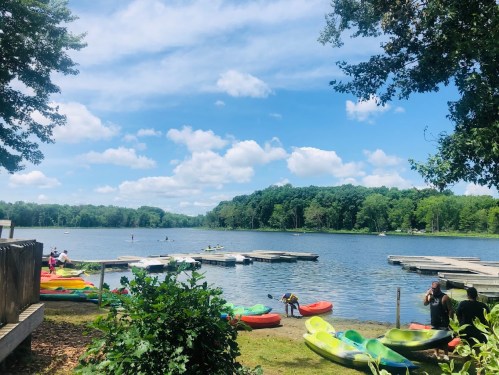 A serene lake scene with colorful kayaks, people paddling, and lush greenery under a bright blue sky with fluffy clouds.