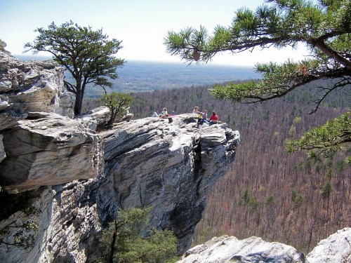 A group of people sitting on a rocky cliff edge, surrounded by trees and a vast landscape in the background.