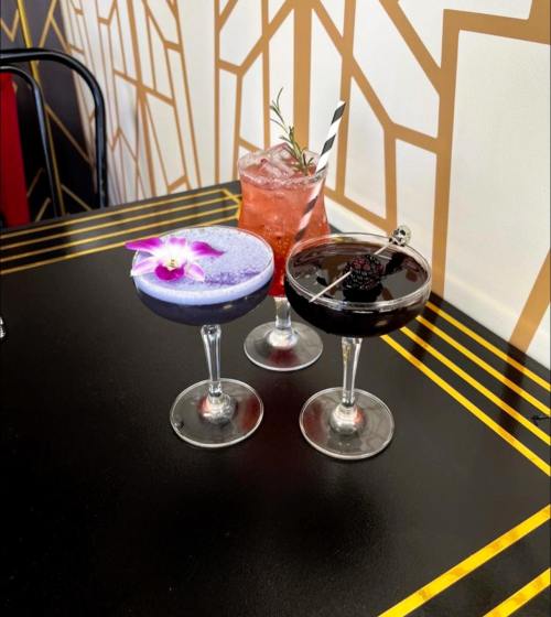 Three colorful cocktails on a table: a purple drink with a flower, a black drink with a berry, and a pink drink with a straw.