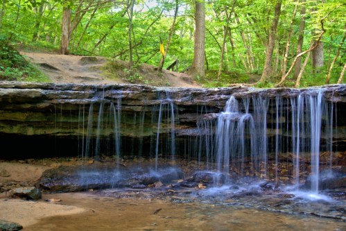 A serene waterfall cascades over rocky ledges, surrounded by lush green trees in a tranquil forest setting.