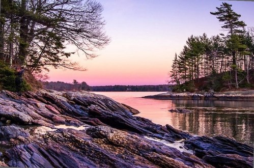 A serene coastal scene at sunset, featuring rocky shores, calm waters, and trees silhouetted against a colorful sky.
