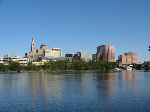 City skyline reflecting on a river, featuring modern buildings and a clear blue sky.