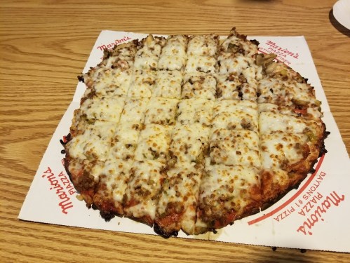 A round pizza with a golden crust, topped with melted cheese and various toppings, cut into square slices on a cardboard tray.