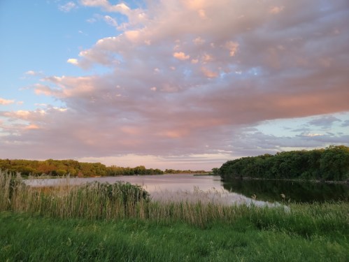 A serene lake surrounded by lush greenery and colorful clouds at sunset, reflecting on the water's surface.