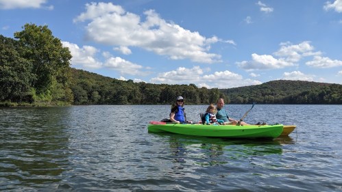A family enjoys kayaking on a calm lake surrounded by green hills and a blue sky with fluffy clouds.