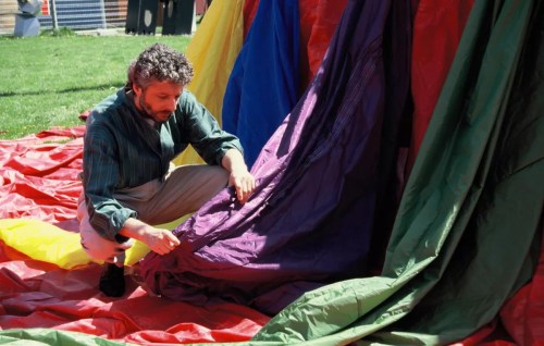 A man adjusts colorful fabric on the ground, surrounded by vibrant red, green, and purple materials in a grassy area.