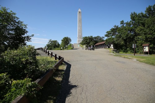 A wide path leads to a tall monument surrounded by trees under a clear blue sky.