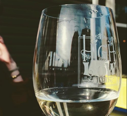 A close-up of a wine glass with the logo of High Low Winery, reflecting a sunny outdoor setting.
