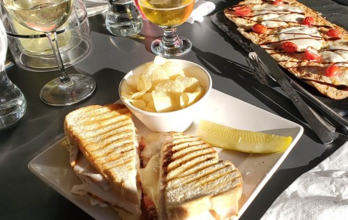 A plate with a grilled sandwich, chips, and a pickle, alongside drinks and a flatbread with tomatoes in the background.