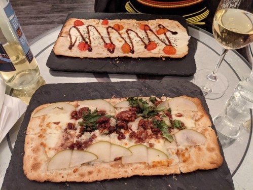Two flatbreads on slate plates: one topped with tomatoes and balsamic, the other with pears, nuts, and herbs, alongside a glass of white wine.