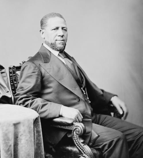 A seated man in formal attire, with a beard, poses confidently in a vintage studio setting.