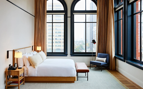 A modern hotel room with a large bed, bench, and armchair, featuring tall windows with city views and warm lighting.
