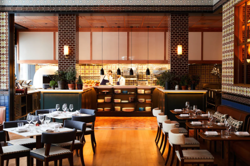 A stylish restaurant interior featuring a kitchen, dining tables, and decorative tiles. Chefs are visible in the background.