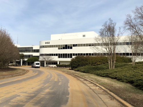 A modern, white building with large windows, surrounded by trees and a winding road leading to its entrance.