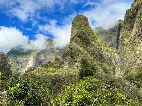 Lush green mountains rise under a blue sky with clouds, showcasing a dramatic landscape.